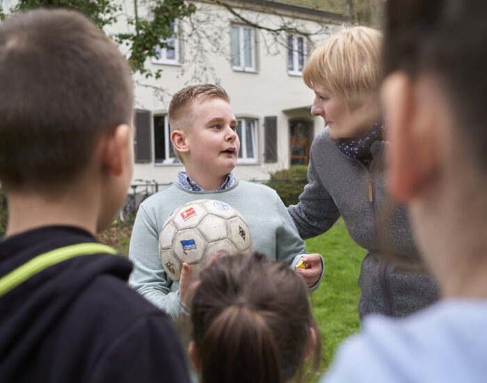 Ein Kind steht vor einer Gruppe Kinder und hält einen Fußball in der Hand. Es wird von einer Schulbegleiterin getröstet.